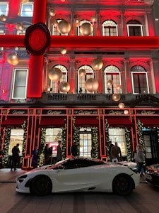 A luxurious storefront is adorned with festive holiday decorations, including large gold ornaments and illuminated garlands. A sleek white sports car is parked in front, with several people casually standing nearby.