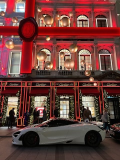 A luxurious storefront is adorned with festive holiday decorations, including large gold ornaments and illuminated garlands. A sleek white sports car is parked in front, with several people casually standing nearby.