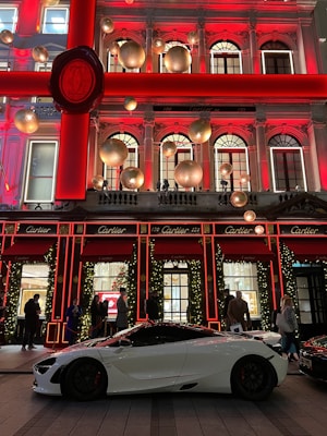 A luxurious storefront is adorned with festive holiday decorations, including large gold ornaments and illuminated garlands. A sleek white sports car is parked in front, with several people casually standing nearby.
