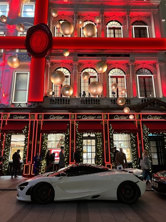 A luxurious storefront is adorned with festive holiday decorations, including large gold ornaments and illuminated garlands. A sleek white sports car is parked in front, with several people casually standing nearby.