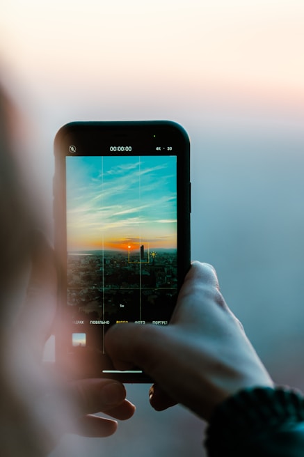 Hands holding a mobile phone capturing a sunset over a city skyline