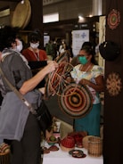People are gathered at a booth displaying handmade woven baskets and crafts. One person is examining a colorful woven item, while the seller, wearing a mask and colorful attire, is standing behind the table filled with similar handcrafted items. The scene appears to be at a marketplace or an exhibition with other booths and partially visible people in the background.