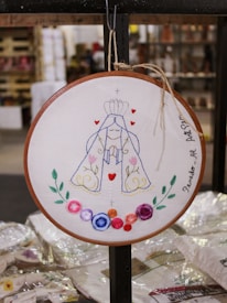 A circular embroidery hoop features an intricately designed embroidered artwork. The central figure appears to be a stylized depiction of a religious icon or saint, adorned with a crown and robe. The design includes elements such as swirling patterns, small hearts, and colorful flowers in various hues surrounding the figure. The embroidery is framed by a simple wooden hoop, and the artwork is titled with writing along the edge.