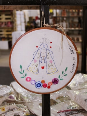 A circular embroidery hoop features an intricately designed embroidered artwork. The central figure appears to be a stylized depiction of a religious icon or saint, adorned with a crown and robe. The design includes elements such as swirling patterns, small hearts, and colorful flowers in various hues surrounding the figure. The embroidery is framed by a simple wooden hoop, and the artwork is titled with writing along the edge.