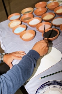 A person is painting on a surface with a brush. Surrounding them are several terracotta bowls filled with different colored pigments, including black, white, blue, and brown. The person is wearing a blue sweater and a watch. A green pencil and a part of a patterned plate are also visible nearby.
