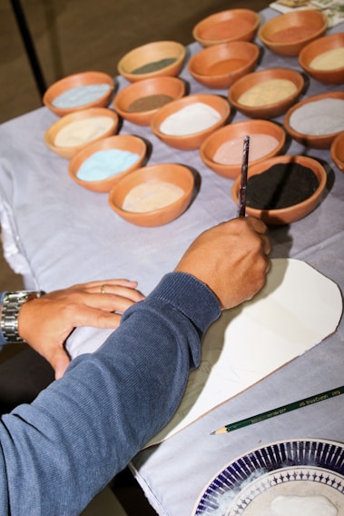 A person is painting on a surface with a brush. Surrounding them are several terracotta bowls filled with different colored pigments, including black, white, blue, and brown. The person is wearing a blue sweater and a watch. A green pencil and a part of a patterned plate are also visible nearby.