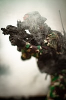 A soldier wearing camouflage gear and holding a rifle is partially obscured by smoke, giving a sense of action or combat. The environment is hazy, suggesting a battlefield or training scenario.