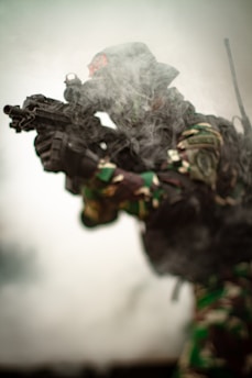 A soldier wearing camouflage gear and holding a rifle is partially obscured by smoke, giving a sense of action or combat. The environment is hazy, suggesting a battlefield or training scenario.