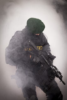 A person wearing black tactical gear and a dark green beret is holding a rifle. The scene is filled with thick white smoke, partially obscuring the individual.
