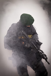 A person wearing black tactical gear and a dark green beret is holding a rifle. The scene is filled with thick white smoke, partially obscuring the individual.
