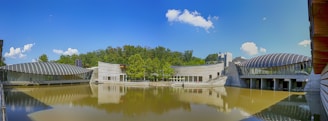 An architectural complex is situated beside a calm body of water, featuring modern buildings with metallic, curved roofs and glass walls. The sky is clear with a few scattered clouds, and lush green trees are in the background, reflecting on the water's surface.
