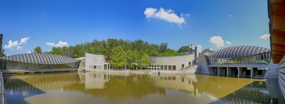 An architectural complex is situated beside a calm body of water, featuring modern buildings with metallic, curved roofs and glass walls. The sky is clear with a few scattered clouds, and lush green trees are in the background, reflecting on the water's surface.