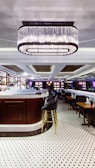An elegant bar interior featuring a large, central bar counter with sleek high chairs surrounding it. Above the bar hangs a prominent, modern chandelier providing ambient lighting. The floor is covered with polished white tiles with black patterns, enhancing the room's sophisticated atmosphere. Shelves behind the counter are stocked with bottles and glasses, adding to the bar-like setting.