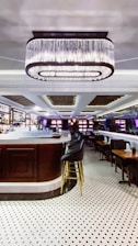 An elegant bar interior featuring a large, central bar counter with sleek high chairs surrounding it. Above the bar hangs a prominent, modern chandelier providing ambient lighting. The floor is covered with polished white tiles with black patterns, enhancing the room's sophisticated atmosphere. Shelves behind the counter are stocked with bottles and glasses, adding to the bar-like setting.