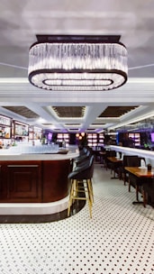 An elegant bar interior featuring a large, central bar counter with sleek high chairs surrounding it. Above the bar hangs a prominent, modern chandelier providing ambient lighting. The floor is covered with polished white tiles with black patterns, enhancing the room's sophisticated atmosphere. Shelves behind the counter are stocked with bottles and glasses, adding to the bar-like setting.