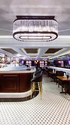 An elegant bar interior featuring a large, central bar counter with sleek high chairs surrounding it. Above the bar hangs a prominent, modern chandelier providing ambient lighting. The floor is covered with polished white tiles with black patterns, enhancing the room's sophisticated atmosphere. Shelves behind the counter are stocked with bottles and glasses, adding to the bar-like setting.