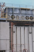 Several large industrial cooling fans are mounted on top of a structure with metal railings and numerous pipes of varying sizes. The fans are enclosed in a horizontal metal frame, and the structure appears to be part of a larger industrial facility. The surface shows signs of wear and weathering.
