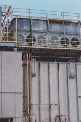 Several large industrial cooling fans are mounted on top of a structure with metal railings and numerous pipes of varying sizes. The fans are enclosed in a horizontal metal frame, and the structure appears to be part of a larger industrial facility. The surface shows signs of wear and weathering.