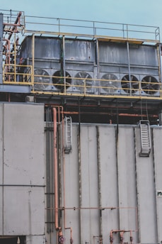 Several large industrial cooling fans are mounted on top of a structure with metal railings and numerous pipes of varying sizes. The fans are enclosed in a horizontal metal frame, and the structure appears to be part of a larger industrial facility. The surface shows signs of wear and weathering.