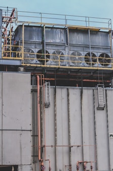 Several large industrial cooling fans are mounted on top of a structure with metal railings and numerous pipes of varying sizes. The fans are enclosed in a horizontal metal frame, and the structure appears to be part of a larger industrial facility. The surface shows signs of wear and weathering.