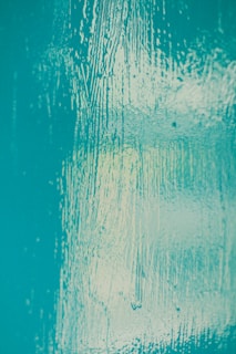 A textured surface with a glossy finish in shades of teal. The surface features visible brush strokes and variations in light reflection, creating a dynamic and visually interesting pattern.