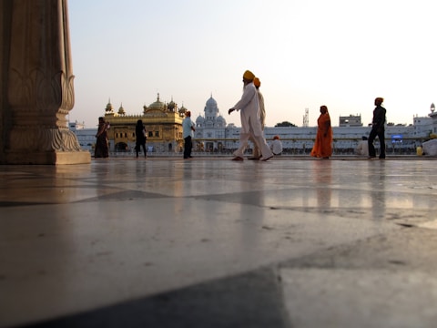 People are walking near a prominent religious structure with golden domes in the background. The ground is made of smooth marble tiles, giving a reflective effect. Several individuals are dressed in traditional attire, including one person wearing a bright orange robe and others donning turbans.