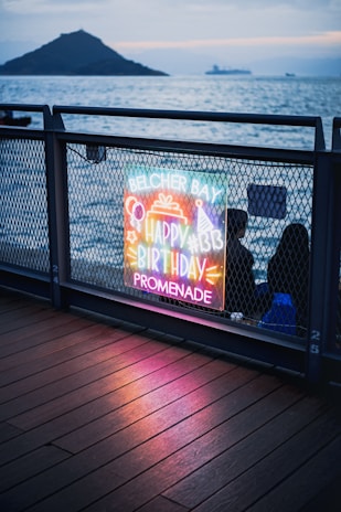 A colorful neon sign with the words 'Belcher Bay Happy Birthday Promenade' is illuminated against a mesh fence. The sign includes decorative symbols like a birthday hat and cake. In the background, there's a view of a calm sea with an island and ships on the horizon, suggesting a waterfront location. The wooden floor beneath reflects the neon lights.