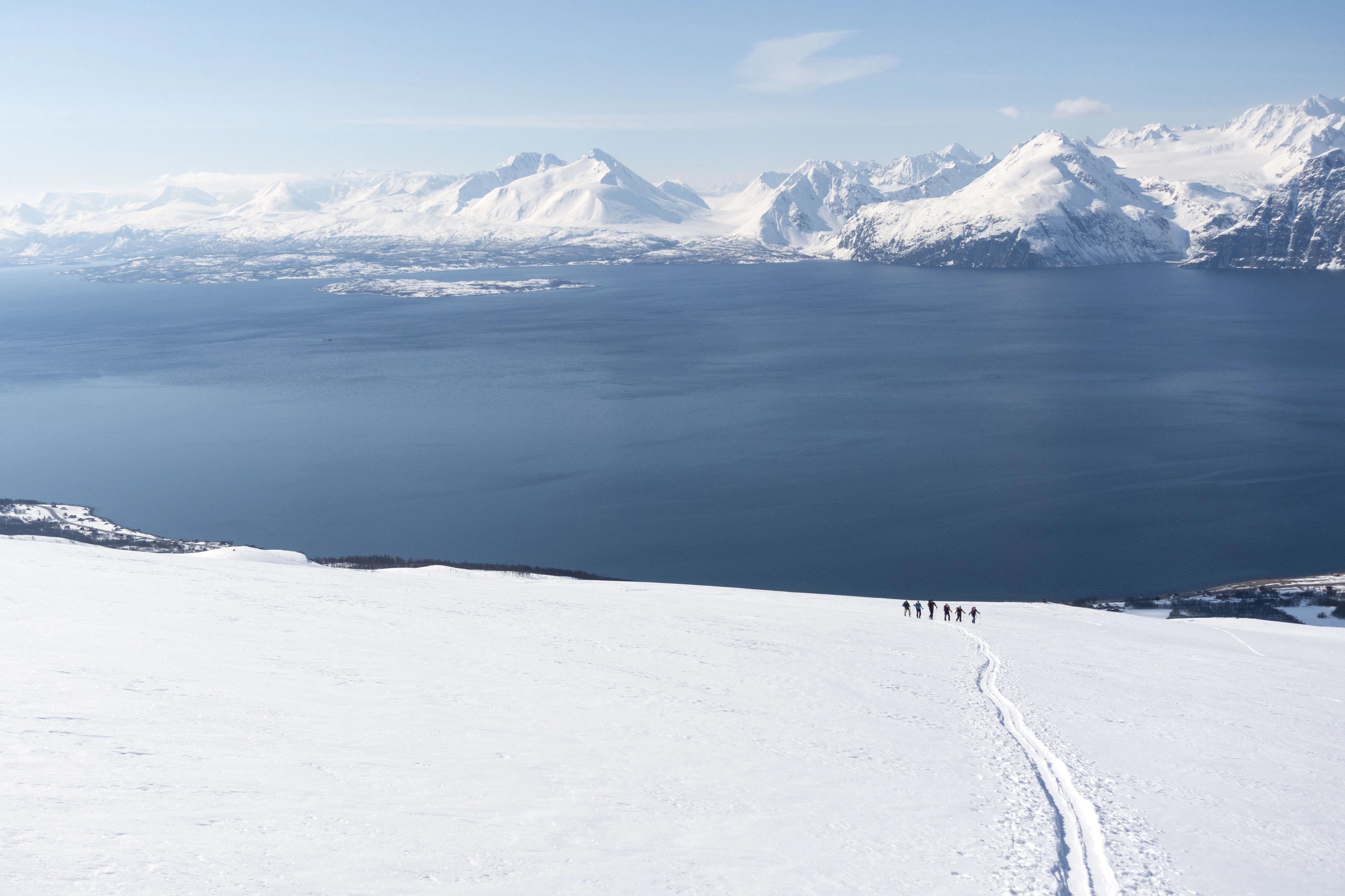 A small group of ski-tourers climbing a snowy slope, with the snow-covered Lyngen Alps across the fjord