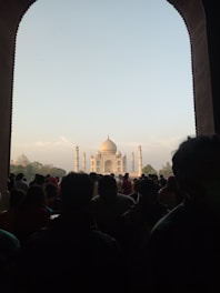 A group of people in silhouette view the iconic white marble mausoleum, the Taj Mahal, framed by a large, dark archway. The sky is clear with light clouds, and natural light illuminates the structure.