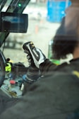 A person is operating machinery from inside a vehicle cabin. The focus is on their hand gripping a joystick-like control device, with various buttons and controls visible on a panel. A screen is mounted in front showing unclear content.