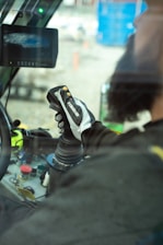 A person is operating machinery from inside a vehicle cabin. The focus is on their hand gripping a joystick-like control device, with various buttons and controls visible on a panel. A screen is mounted in front showing unclear content.