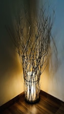A warm light fixture is surrounded by an arrangement of slender branches placed in a corner. The softly glowing lamp creates intricate shadows on the walls, enhancing the rustic and cozy atmosphere. The floor has a wooden texture, adding to the earthy and natural aesthetic.