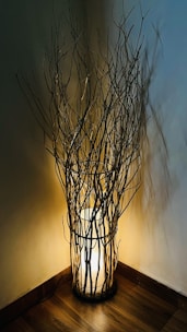 A warm light fixture is surrounded by an arrangement of slender branches placed in a corner. The softly glowing lamp creates intricate shadows on the walls, enhancing the rustic and cozy atmosphere. The floor has a wooden texture, adding to the earthy and natural aesthetic.