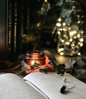 A cozy scene featuring an open journal with handwritten notes, a lit candle in a glass jar casting a warm glow, and a string of fairy lights inside a transparent container. Green leaves and a black crystal pendant on a silver chain are also present, enhancing the serene setting.