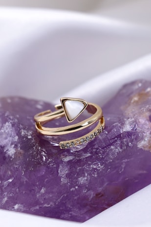 A gold ring with a triangular white centerpiece and small gemstones set in a curve rests on a purple crystalline surface.