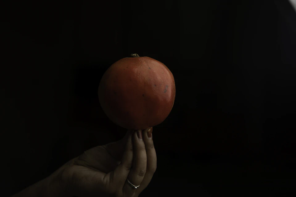The image shows a hand holding up a single, vibrant red pomegranate against a stark black background. The pomegranate is positioned centrally within the frame and appears to be the main subject of the photograph.