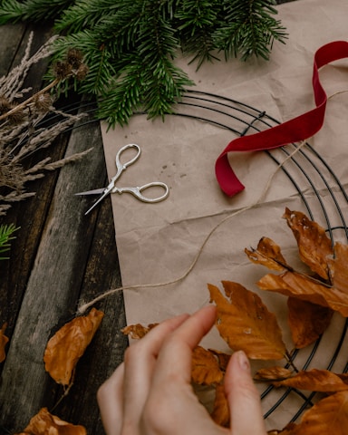 A rustic scene featuring a wire wreath frame with brown leaves and evergreen sprigs on a wooden table. A pair of silver scissors lies nearby, with a red ribbon and a piece of twine. A hand is reaching towards the autumn leaves, suggesting a crafting activity.