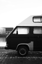 A black and white photograph of a parked van with a high roof, positioned near a body of water. The vehicle is captured from the side, showcasing the detailed texture of its metal body, and condensation on the windows. The setting appears industrial, with warehouses reflected in the background water, and a cobblestone path in the foreground.