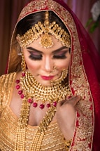 A woman is adorned in elaborate traditional jewelry, including a gold necklace, earrings, and headpiece, featuring intricate designs with red gemstones. She is wearing a richly embroidered red and gold veil and attire. Her makeup is glamorous with bold eyelashes and bright lipstick, creating an elegant appearance.