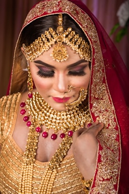 A woman is adorned in elaborate traditional jewelry, including a gold necklace, earrings, and headpiece, featuring intricate designs with red gemstones. She is wearing a richly embroidered red and gold veil and attire. Her makeup is glamorous with bold eyelashes and bright lipstick, creating an elegant appearance.