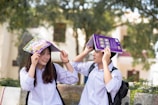 Two students wearing white uniforms are sitting on a bench outdoors, holding books over their heads playfully. The background features trees and a blurred building, suggesting a campus setting. Both students are smiling and appear to be enjoying a lighthearted moment.