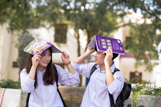 Two students wearing white uniforms are sitting on a bench outdoors, holding books over their heads playfully. The background features trees and a blurred building, suggesting a campus setting. Both students are smiling and appear to be enjoying a lighthearted moment.