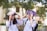 Two students wearing white uniforms are sitting on a bench outdoors, holding books over their heads playfully. The background features trees and a blurred building, suggesting a campus setting. Both students are smiling and appear to be enjoying a lighthearted moment.