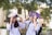 Two students wearing white uniforms are sitting on a bench outdoors, holding books over their heads playfully. The background features trees and a blurred building, suggesting a campus setting. Both students are smiling and appear to be enjoying a lighthearted moment.