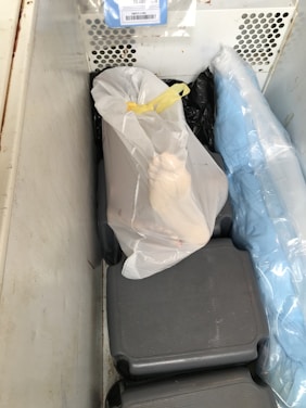 A tightly packed freezer contains several objects wrapped in plastic bags. One of the bags is white with a black lining and tied with a yellow tie. A gloved hand is visible resting on the package. The surrounding bags have varying shades, including blue.