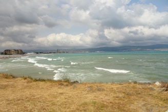 The image features a scenic view of a coastline with a beach and turquoise sea waves gently reaching the shore. In the distance, there are buildings and a small cityscape under a sky filled with dramatic, cloudy skies. The foreground is characterized by dry, yellowish grass and some rocks.