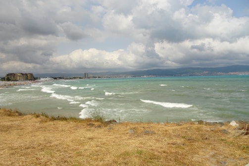 The image features a scenic view of a coastline with a beach and turquoise sea waves gently reaching the shore. In the distance, there are buildings and a small cityscape under a sky filled with dramatic, cloudy skies. The foreground is characterized by dry, yellowish grass and some rocks.