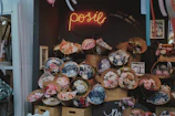A flower shop display featuring numerous bouquets of flowers in various colors and sizes arranged in front of a dark wall. The shop has a warm and cozy atmosphere, with a neon sign that reads 'posie' and a small framed picture on the wall.