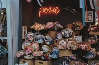A flower shop display featuring numerous bouquets of flowers in various colors and sizes arranged in front of a dark wall. The shop has a warm and cozy atmosphere, with a neon sign that reads 'posie' and a small framed picture on the wall.