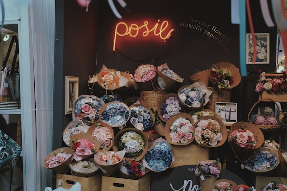 A flower shop display featuring numerous bouquets of flowers in various colors and sizes arranged in front of a dark wall. The shop has a warm and cozy atmosphere, with a neon sign that reads 'posie' and a small framed picture on the wall.